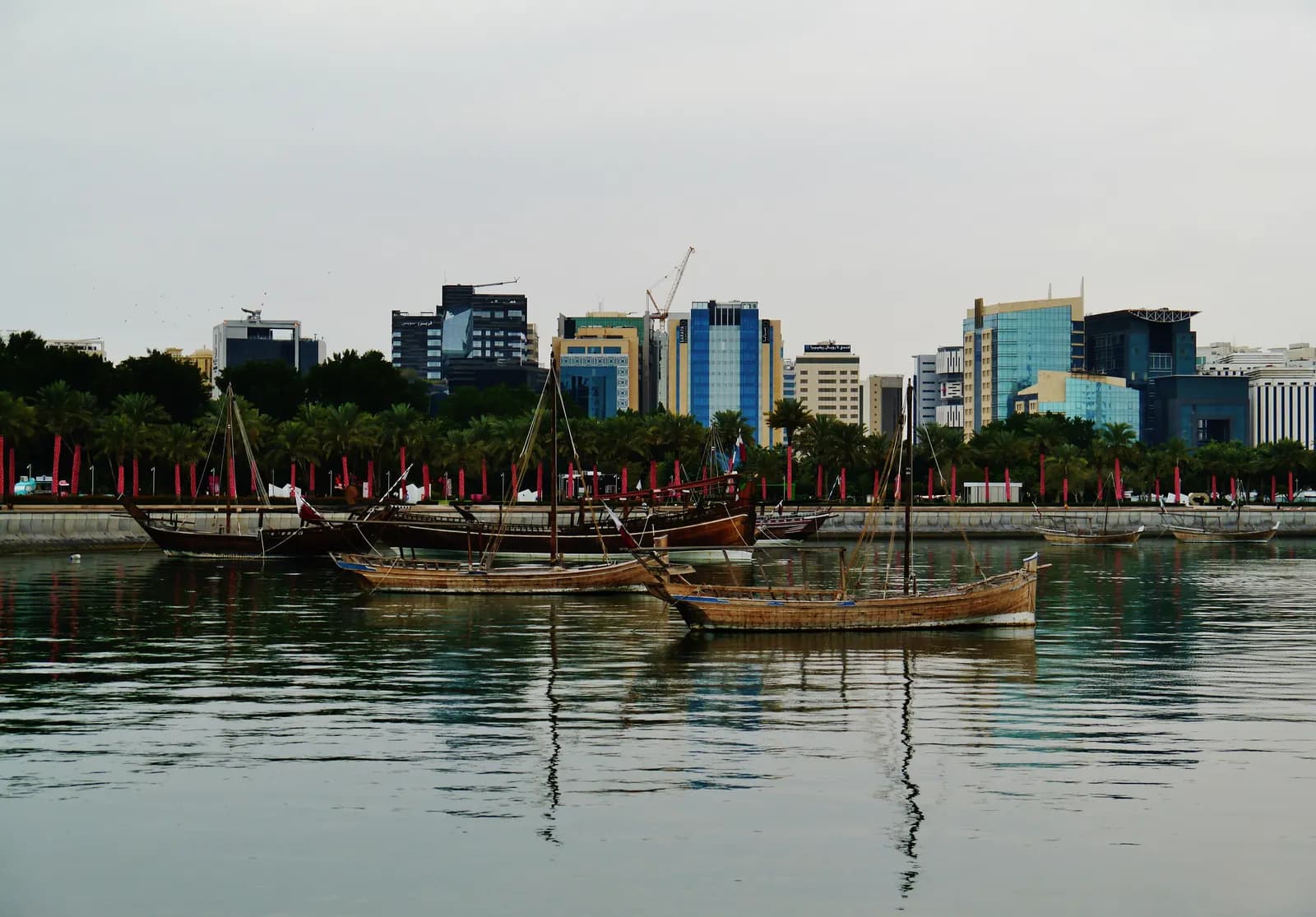 Wooden dhows moored in Doha Bay off the Corniche, seen from MIA Park, with palm trees along the waterfront and office buildings behind
