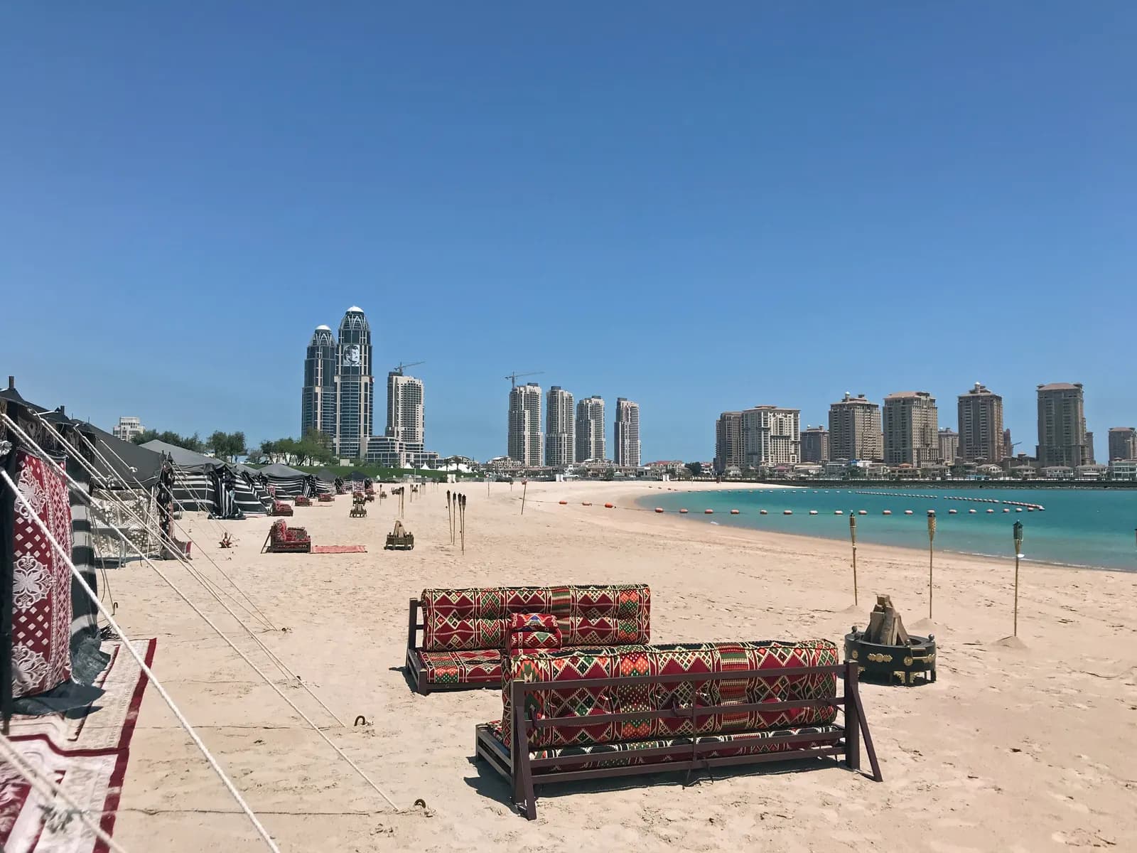 Katara Beach with patterned floor cushions, tents and torches on the sand, a buoy line in turquoise water and residential towers across the bay