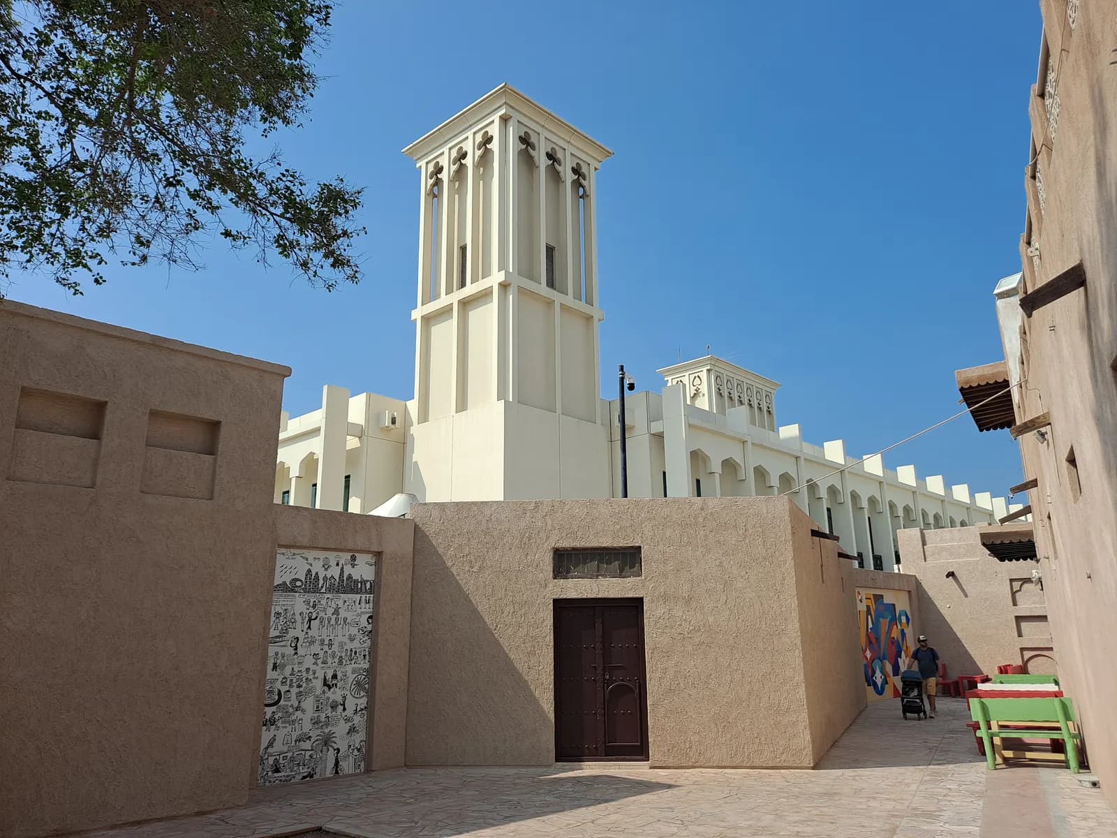 A wind tower rising above sand-coloured walls and a wooden door in the Al Fahidi Historical Neighbourhood of Bur Dubai, with murals on the walls