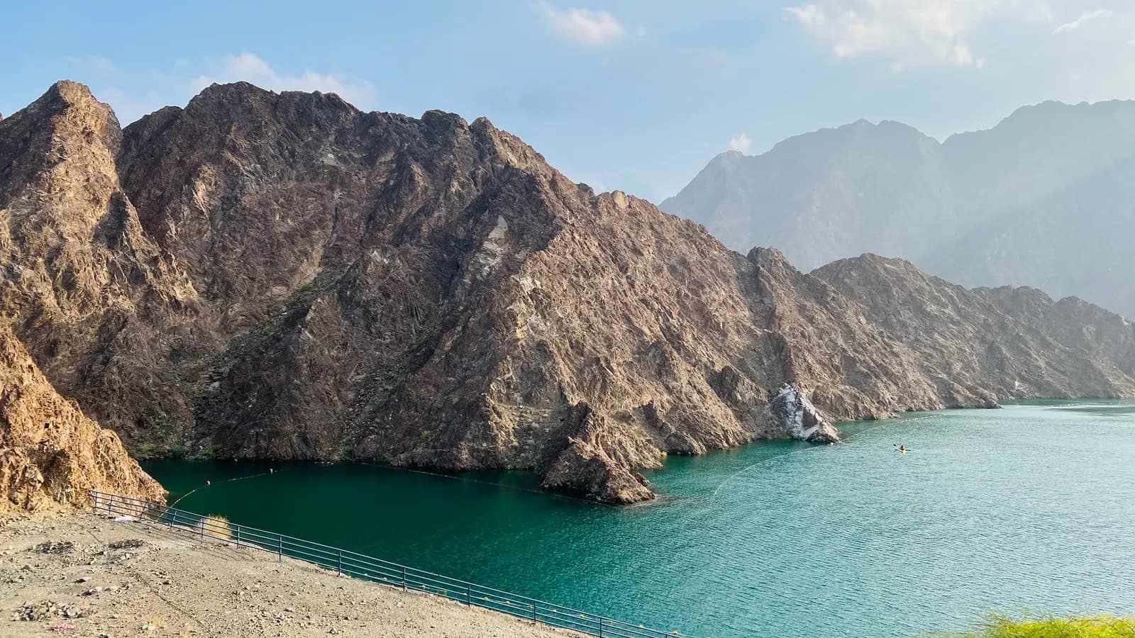 The turquoise reservoir of Hatta Dam below bare, rocky mountain slopes, with a fence along the shore