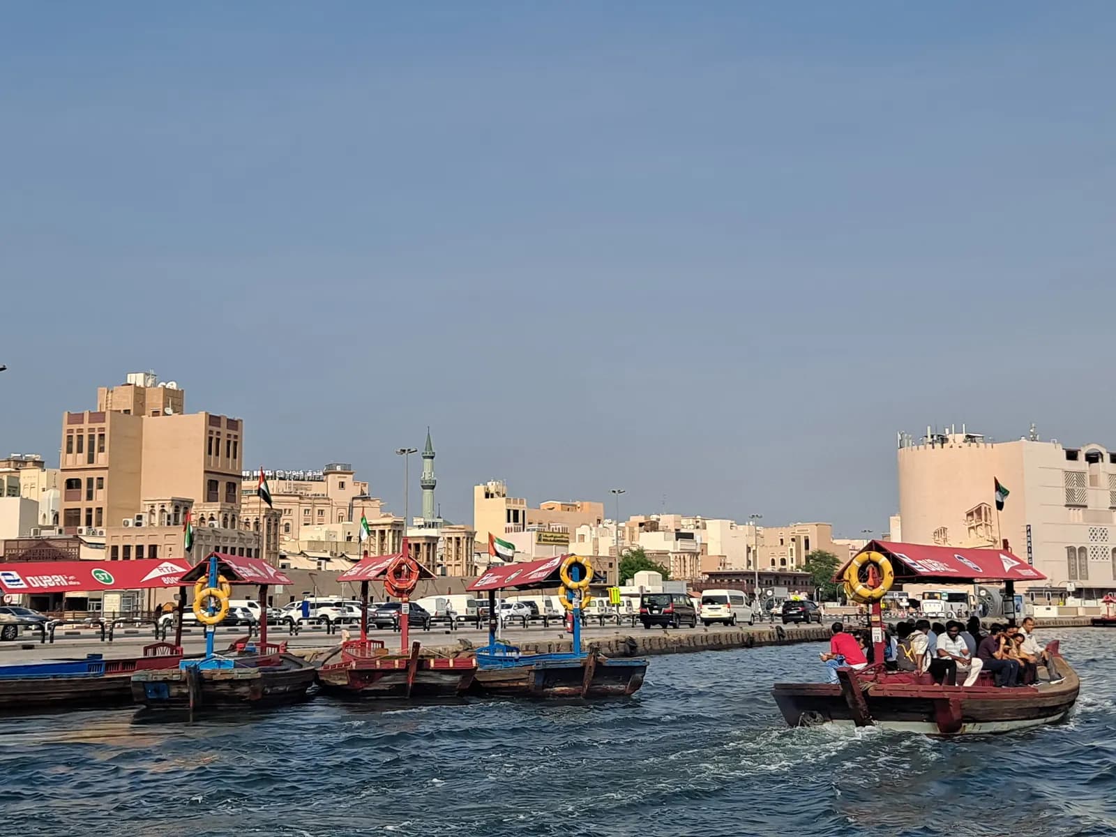 Abra water taxis with red canopies on Dubai Creek, one crossing with passengers, and low buildings with a minaret along the waterfront behind