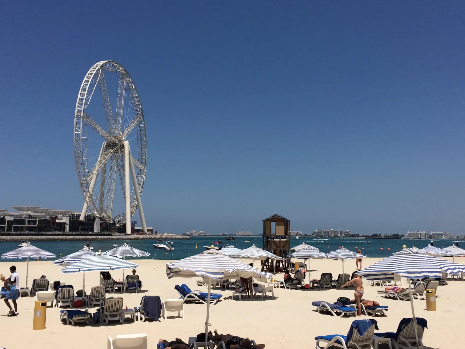 Striped umbrellas and sun loungers on the beach at JBR, with a large observation wheel standing across the water