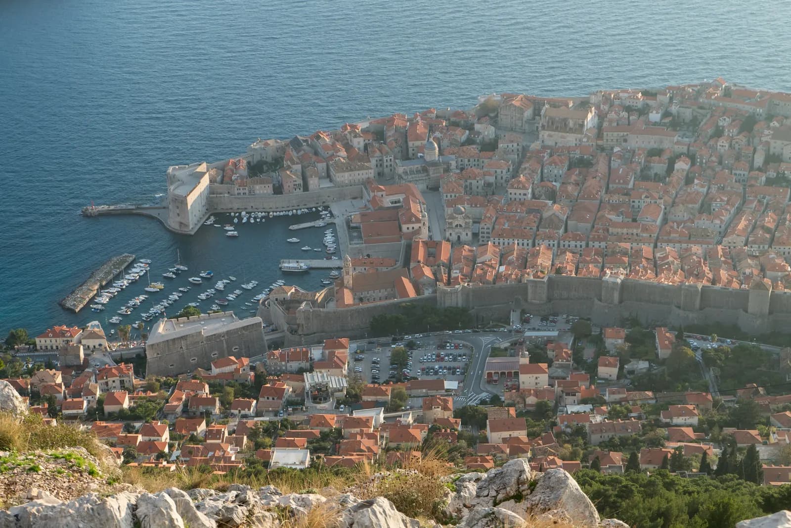 Dubrovnik's walled Old Town and old harbour seen from Mount Srđ, with the red roofs of the Ploče district and a car park below the city walls