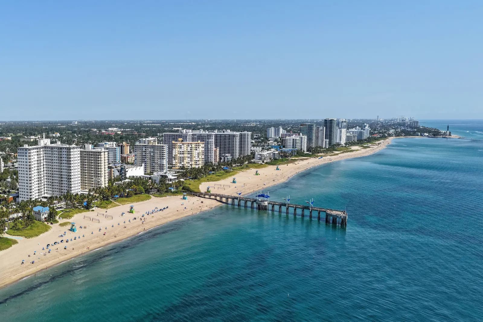 Aerial view of Pompano Beach, with apartment towers behind a wide sandy beach, a long pier reaching into the turquoise sea and a lighthouse at the inlet in the far distance