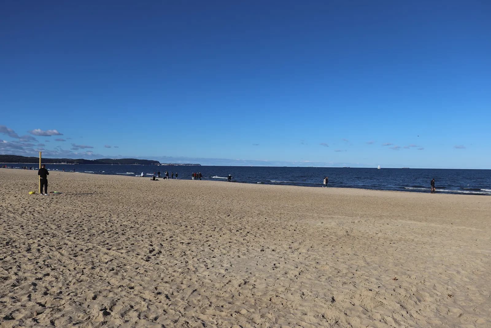 The wide sandy beach at Jelitkowo in Gdańsk under a clear blue sky, with a few walkers at the water's edge