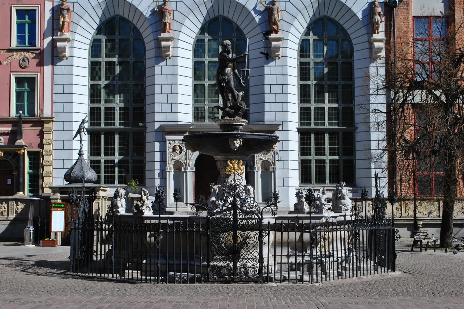 The bronze Neptune fountain behind a wrought iron railing on Długi Targ in Gdańsk, in front of a pale facade with tall arched windows