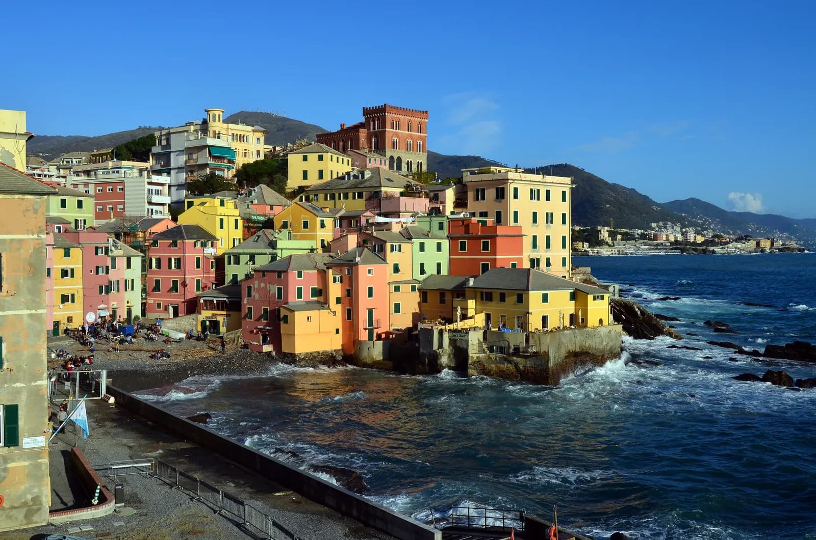 The old fishing cove of Boccadasse in Genoa: pink, yellow and green houses stacked above a pebble beach, with surf breaking on the rocks and the coast running east.
