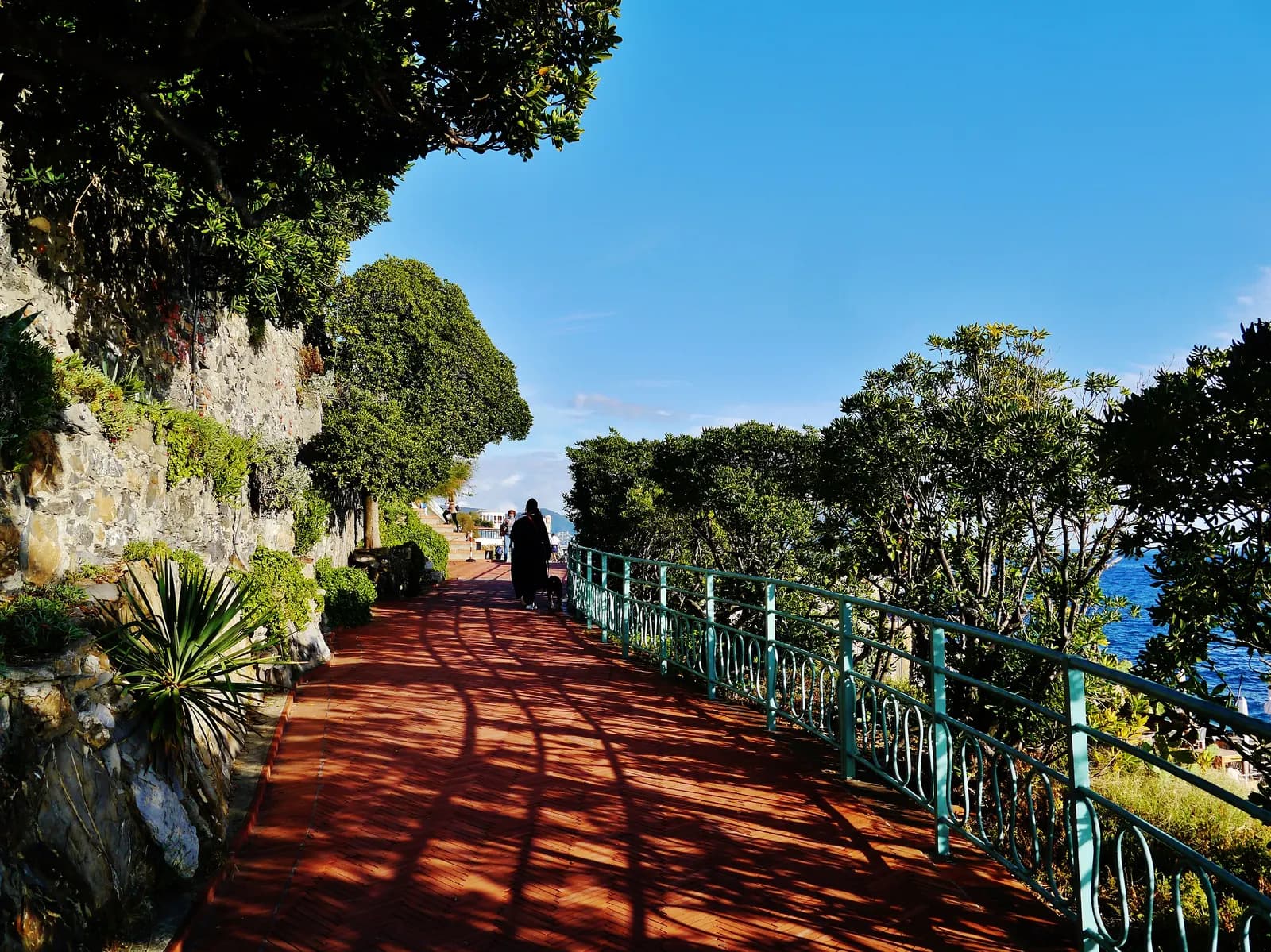 The red brick Passeggiata Anita Garibaldi at Nervi in Genoa, a paved clifftop walk with a green railing, shrubs and the sea on the right.