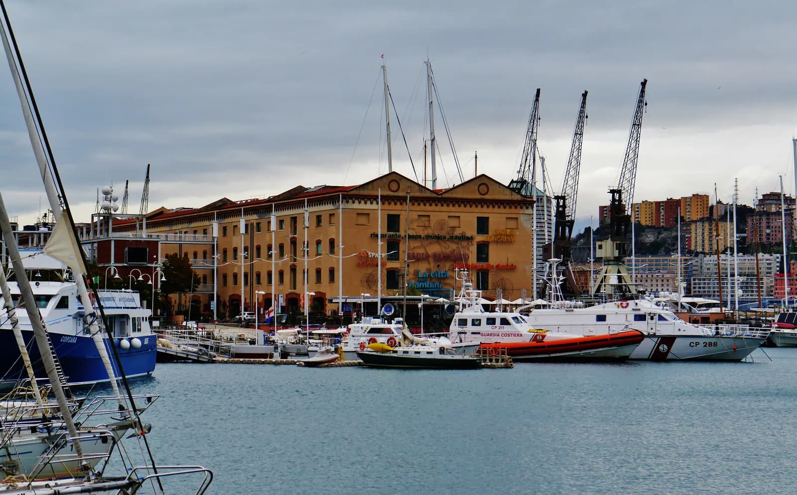 The Porto Antico in Genoa under a grey sky: moored yachts and a coastguard boat in front of the long ochre Magazzini del Cotone building and harbour cranes.