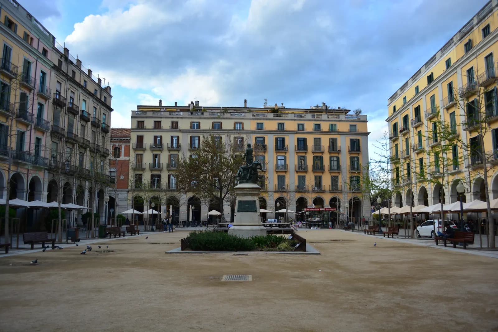 Plaça de la Independència in Girona, an arcaded square of pale yellow buildings with café terraces and a monument at its centre.