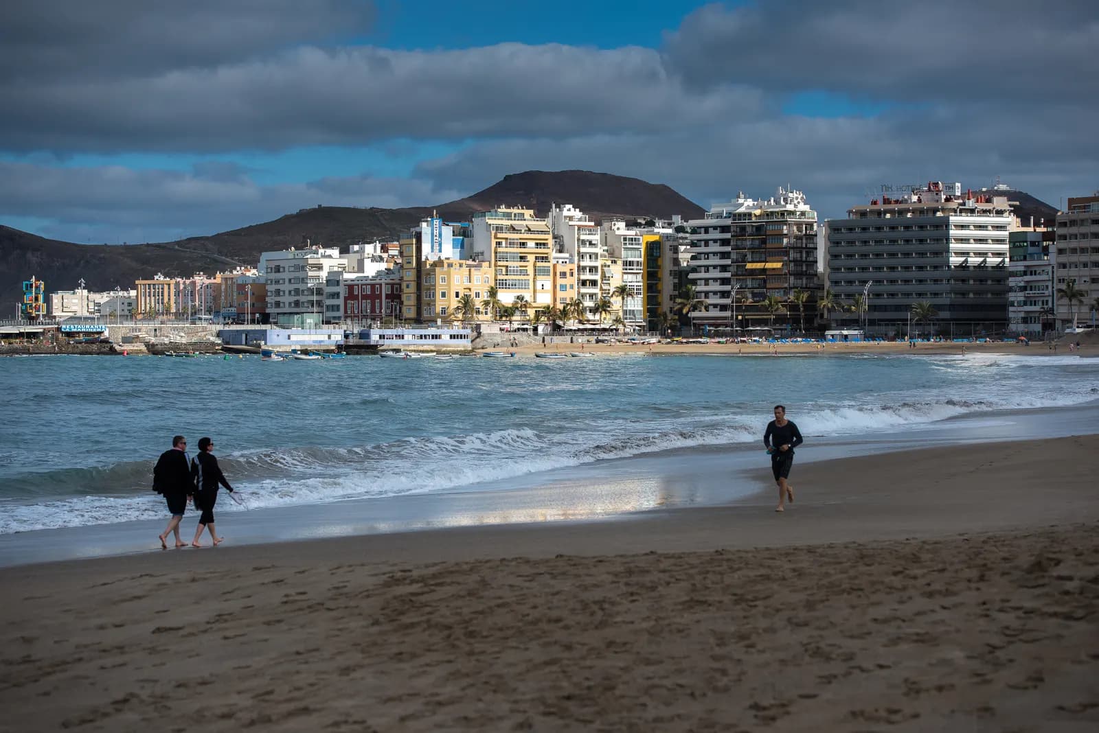 People walking along the sand of Las Canteras beach with apartment blocks and hotels on the seafront and hills behind