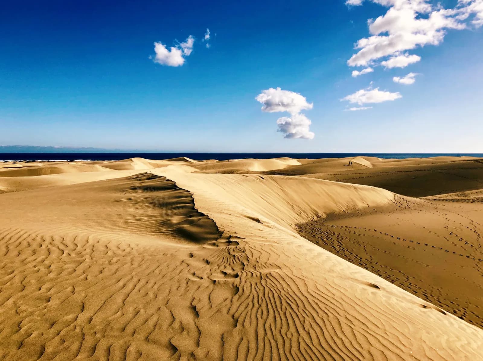 Rippled sand dunes of Maspalomas under a blue sky with a few clouds, with the sea on the horizon