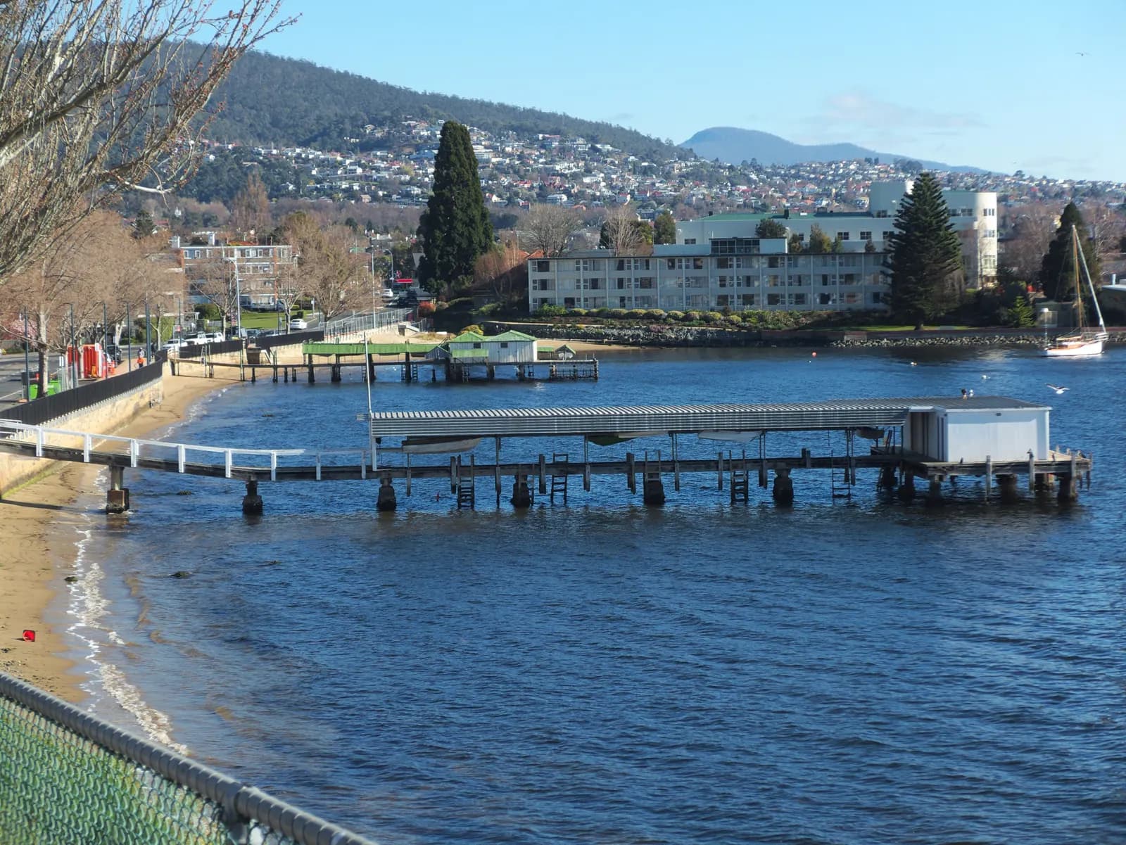Lords Beach in Sandy Bay, Hobart, with two covered jetties over the water, an apartment building among trees and houses on the hills behind