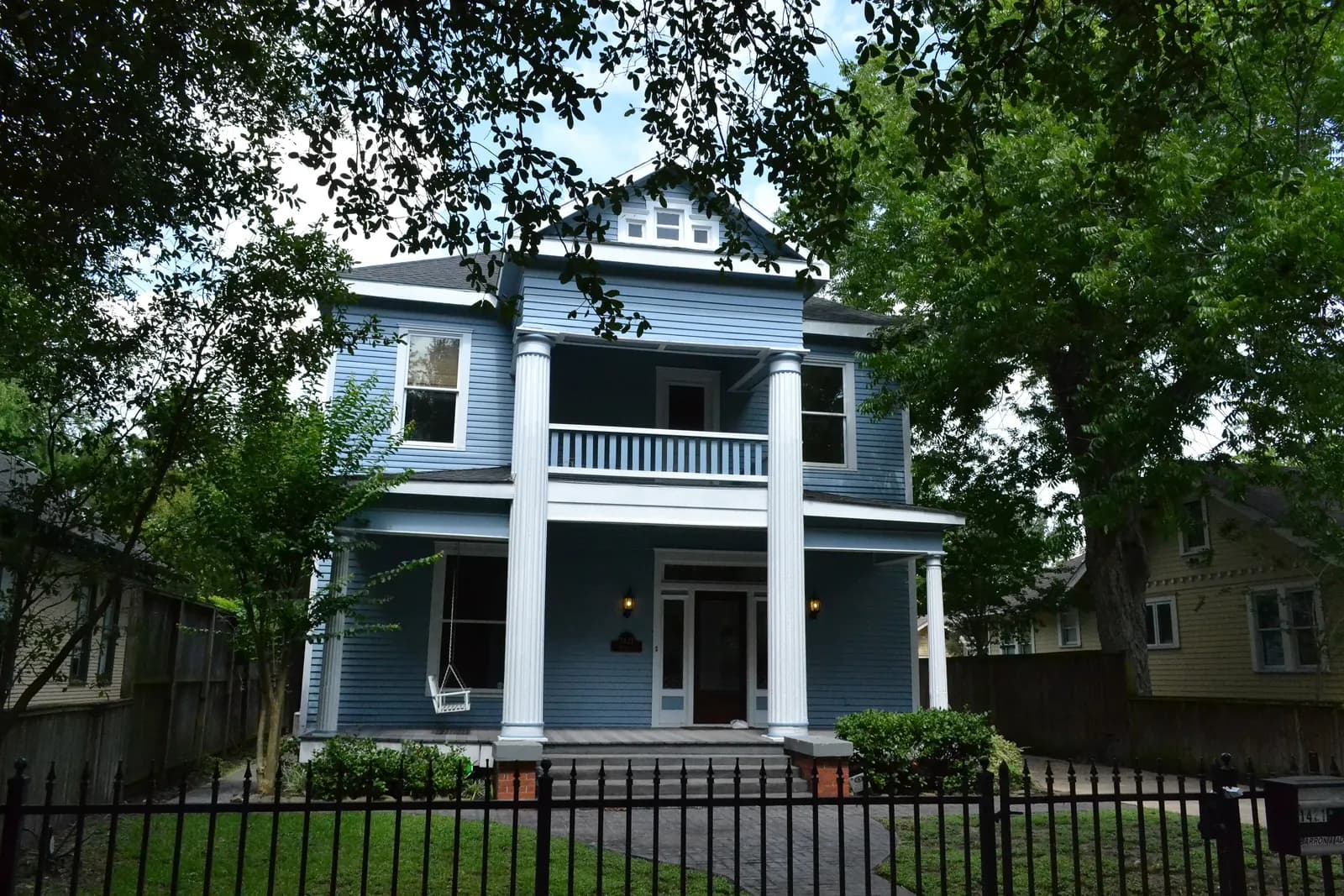 A blue two-storey wooden house with white columns and a porch swing behind an iron fence on Heights Boulevard, Houston