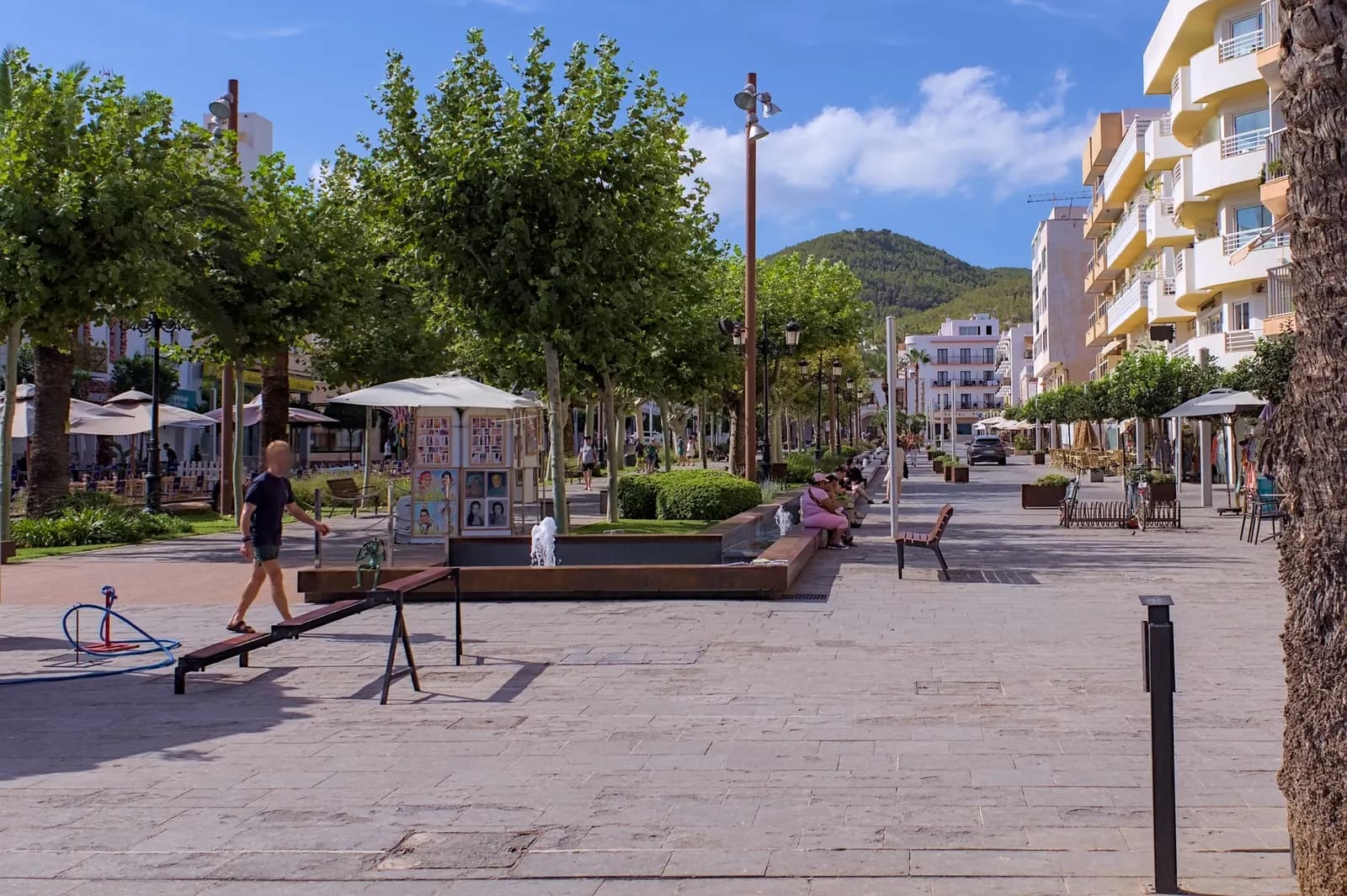 The Passeig de s'Alamera in Santa Eulària des Riu, a paved promenade with plane trees, a fountain and apartment buildings, below a wooded hill.