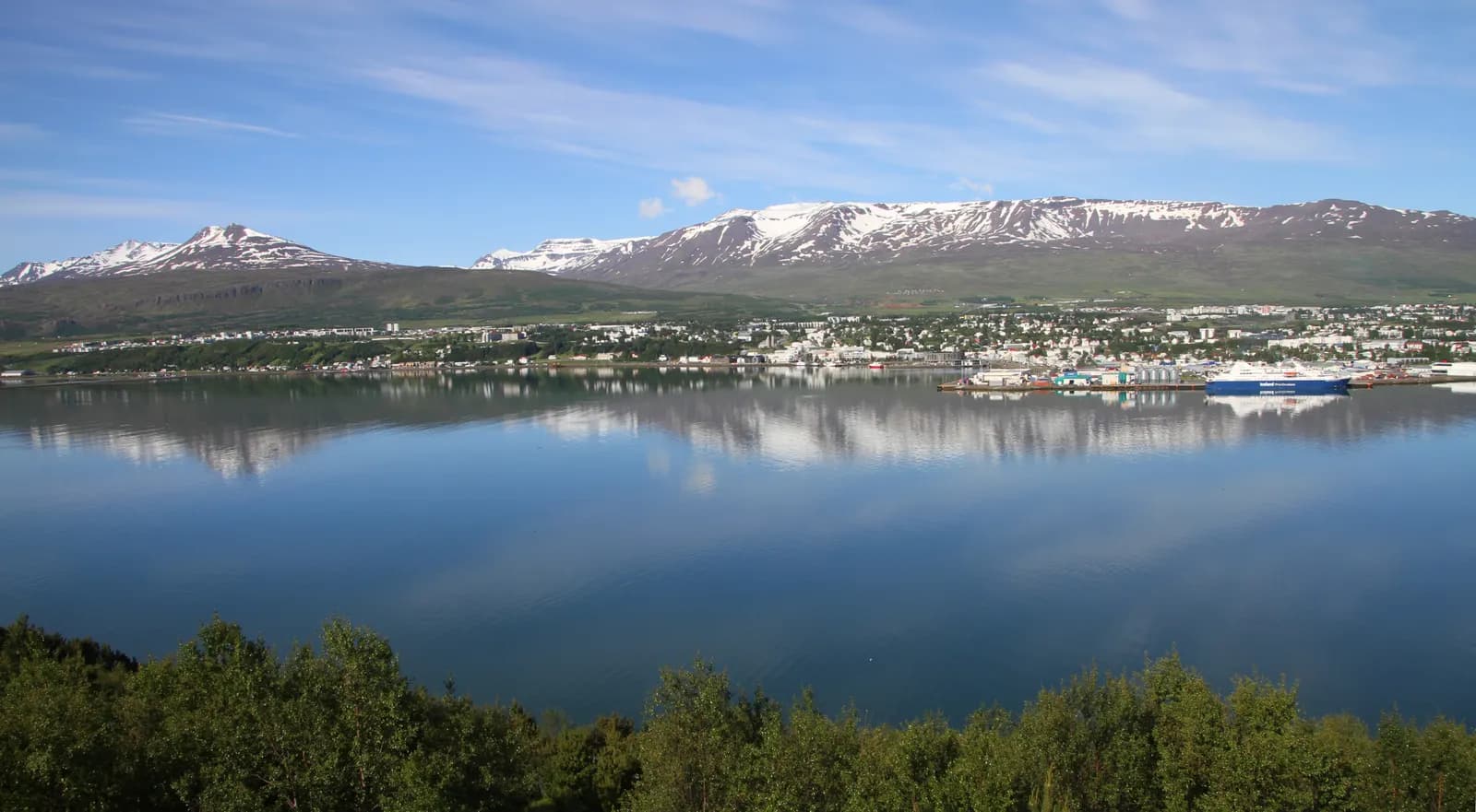 Akureyri seen across the calm water of Eyjafjörður, the town reflected below snow-streaked mountains, with trees in the foreground
