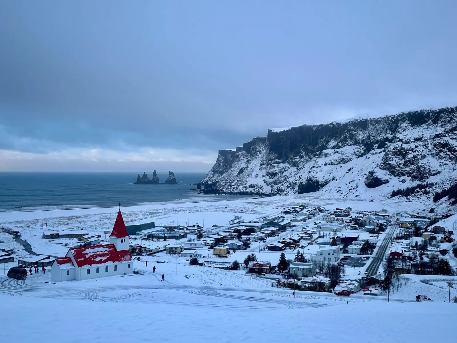 The village of Vík í Mýrdal under snow, with its red-roofed church on the slope, the black beach, the Reynisdrangar sea stacks and the cliffs of Reynisfjall behind