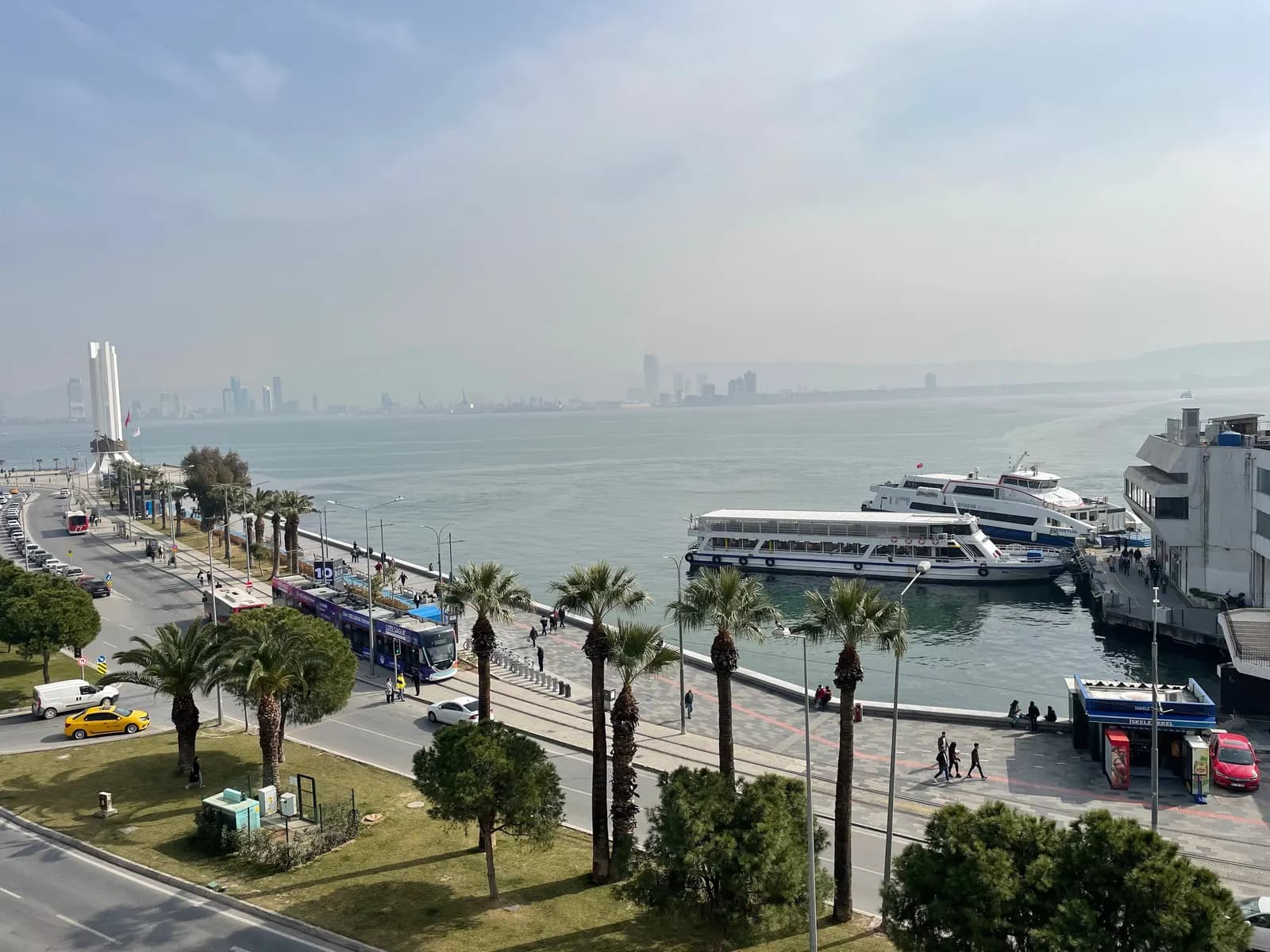 City ferries at the Karşıyaka pier, with a tram on the palm-lined seafront road and the towers of the city across the bay