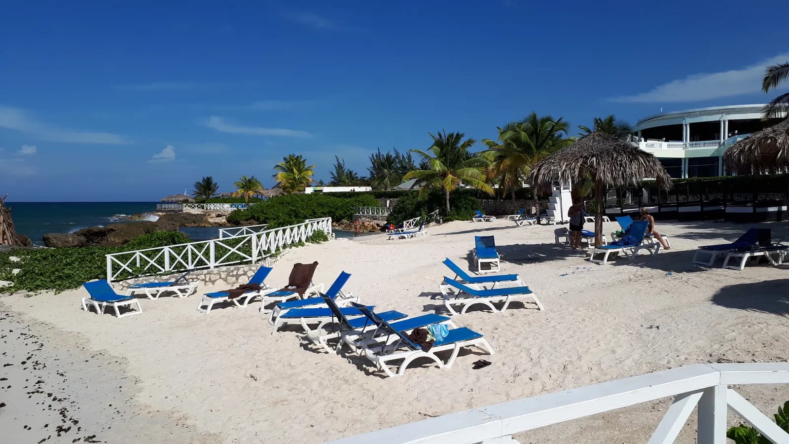 Blue sun loungers on white sand at a beach in Montego Bay, with a white railing, palm trees, a thatched shade and a rocky shore