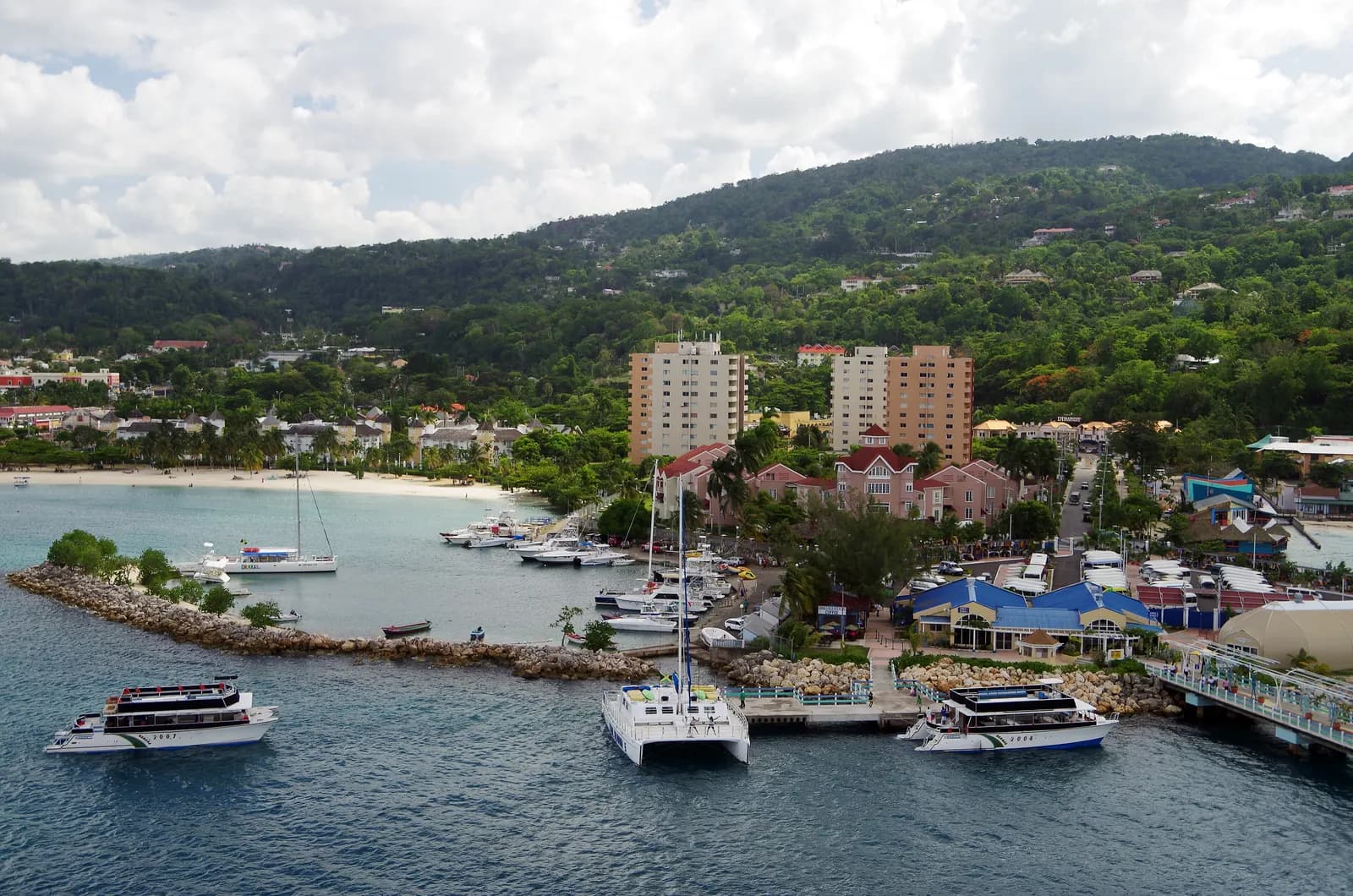 Ocho Rios seen from the sea, with tour boats at a jetty, a small beach and marina, apartment towers and green hills behind