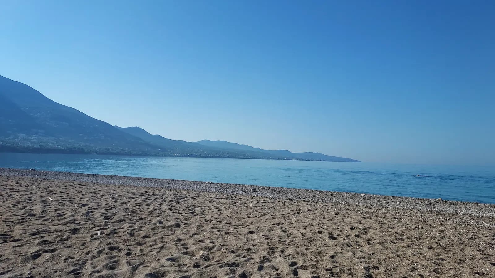 The long sand and pebble beach on the east side of Kalamata, with calm water of the Messinian Gulf and mountains along the coast in the haze