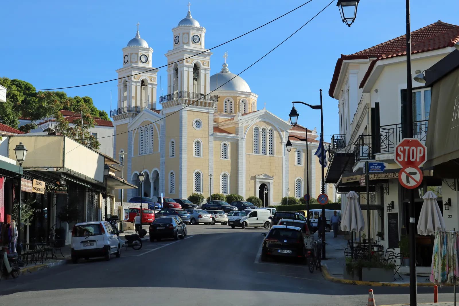 The Ypapanti church in Kalamata with two clock towers and a dome at the end of a street lined with parked cars, cafes and shops under a clear sky