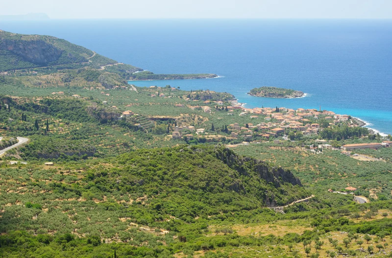 The village of Kardamyli seen from the hills above, red roofs among olive groves by a turquoise sea, with a small wooded islet offshore and the coast of the Mani beyond