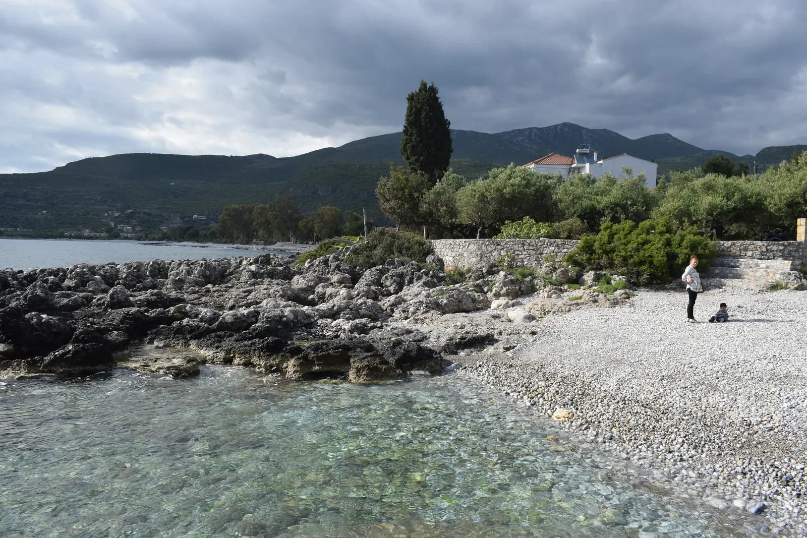 A rocky shore and pebble beach at Kardamyli with clear shallow water, a stone wall, olive trees, a cypress and a white house under a cloudy sky
