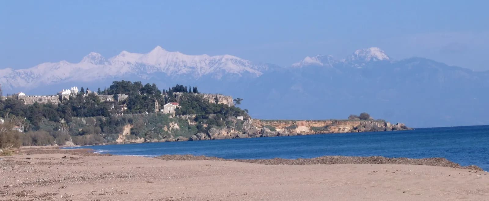 Koroni on its headland above the sea, with the castle walls and houses among trees, seen across a sandy beach with snow-covered mountains behind