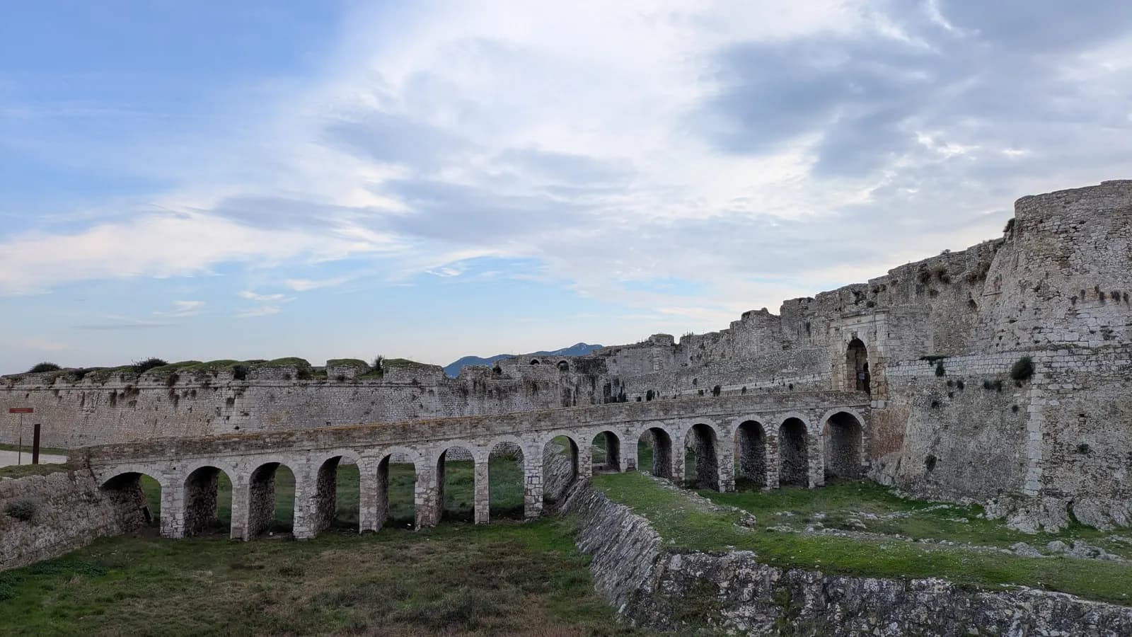 The arched stone bridge across the dry moat to the gate of the castle at Methoni, with the long fortified walls and a pale sky