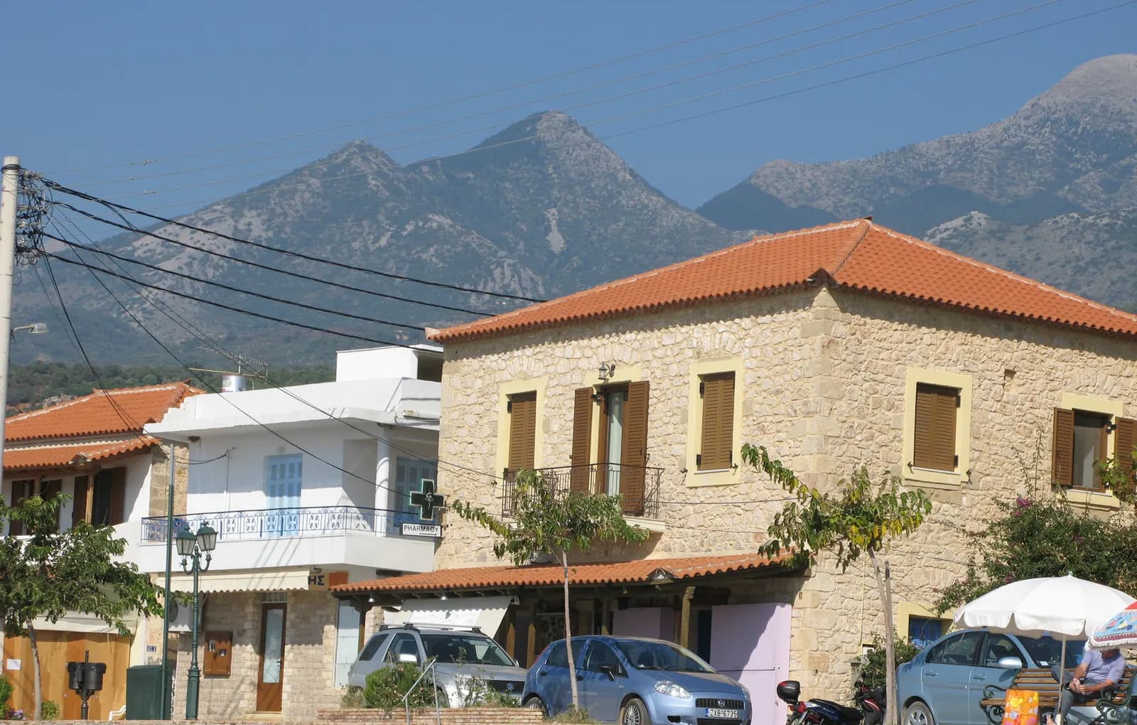 Stone and white houses with tiled roofs behind the beach at Stoupa, parked cars and a beach umbrella in front, and bare mountain peaks behind