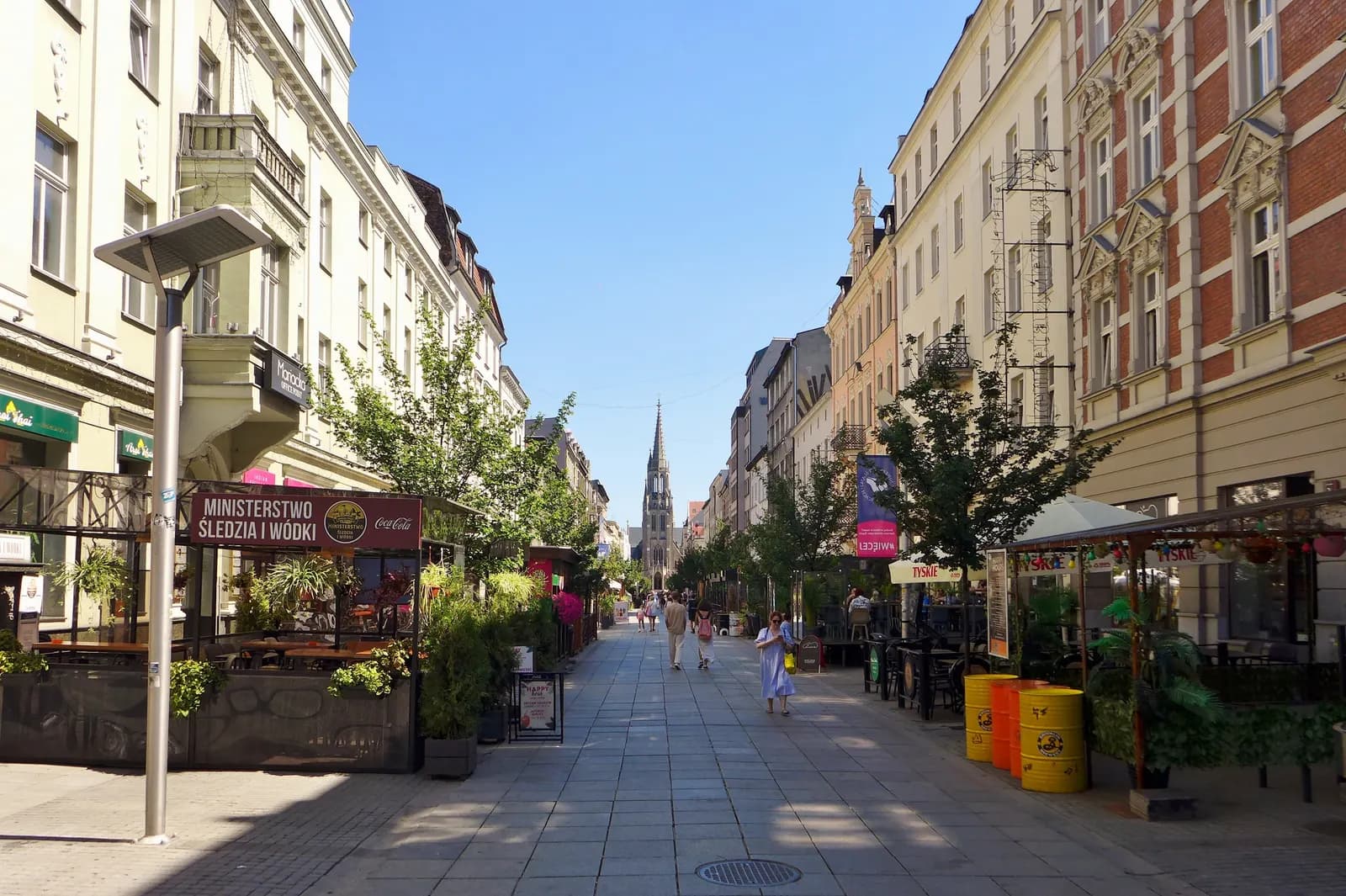 Mariacka Street in central Katowice, a pedestrian street of tall tenement houses with café terraces and trees, leading to the spire of St Mary's Church