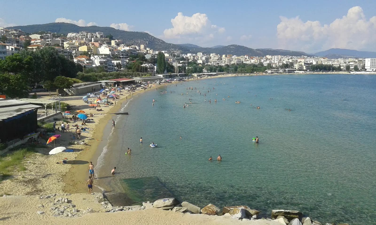 The sandy beach of Kalamitsa on the west side of Kavala, with swimmers in clear shallow water, parasols on the sand and apartment blocks on the hills behind