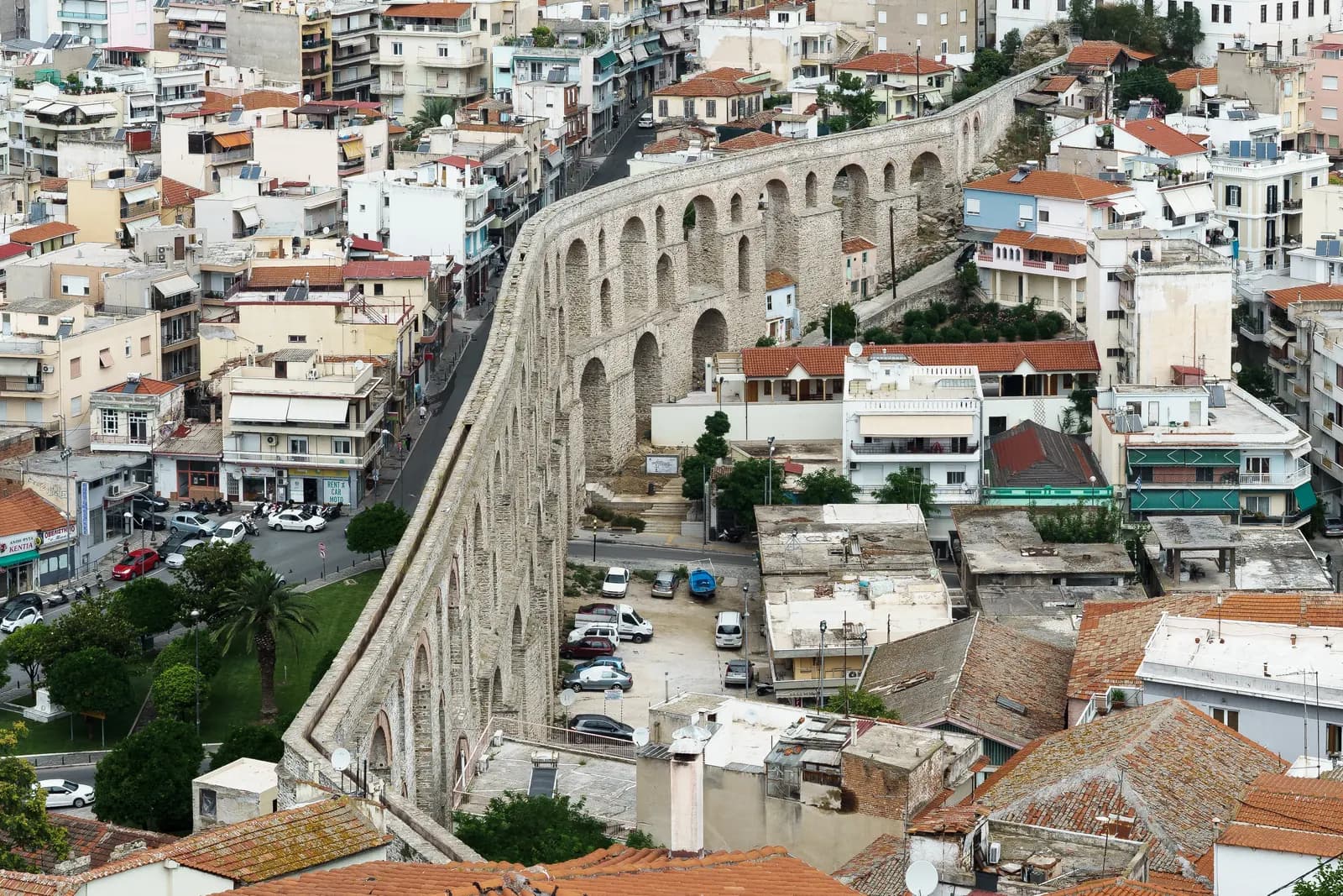 The stone arches of the Kamares aqueduct curving through central Kavala between apartment blocks, roofs and a car park, seen from above