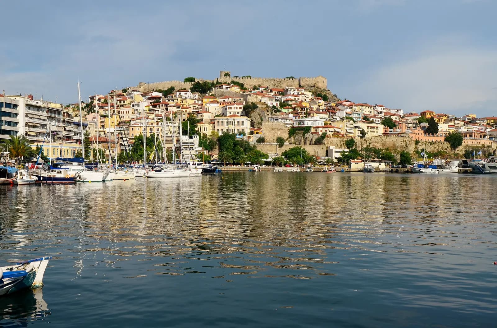The old town of Kavala climbing the Panagia peninsula to the castle walls, with sailing boats in the marina and the houses reflected in calm water