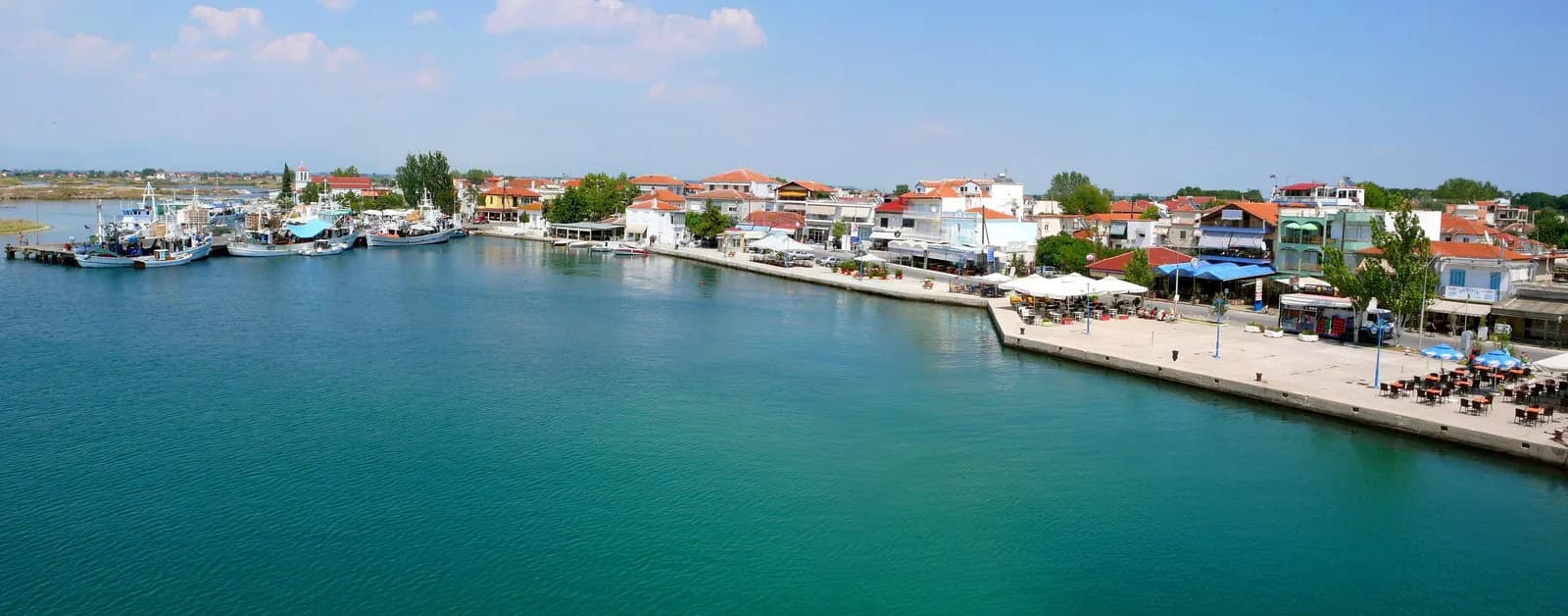 The harbour front of Keramoti with fishing boats moored on the left, cafe terraces along the quay and low houses with tiled roofs under a summer sky