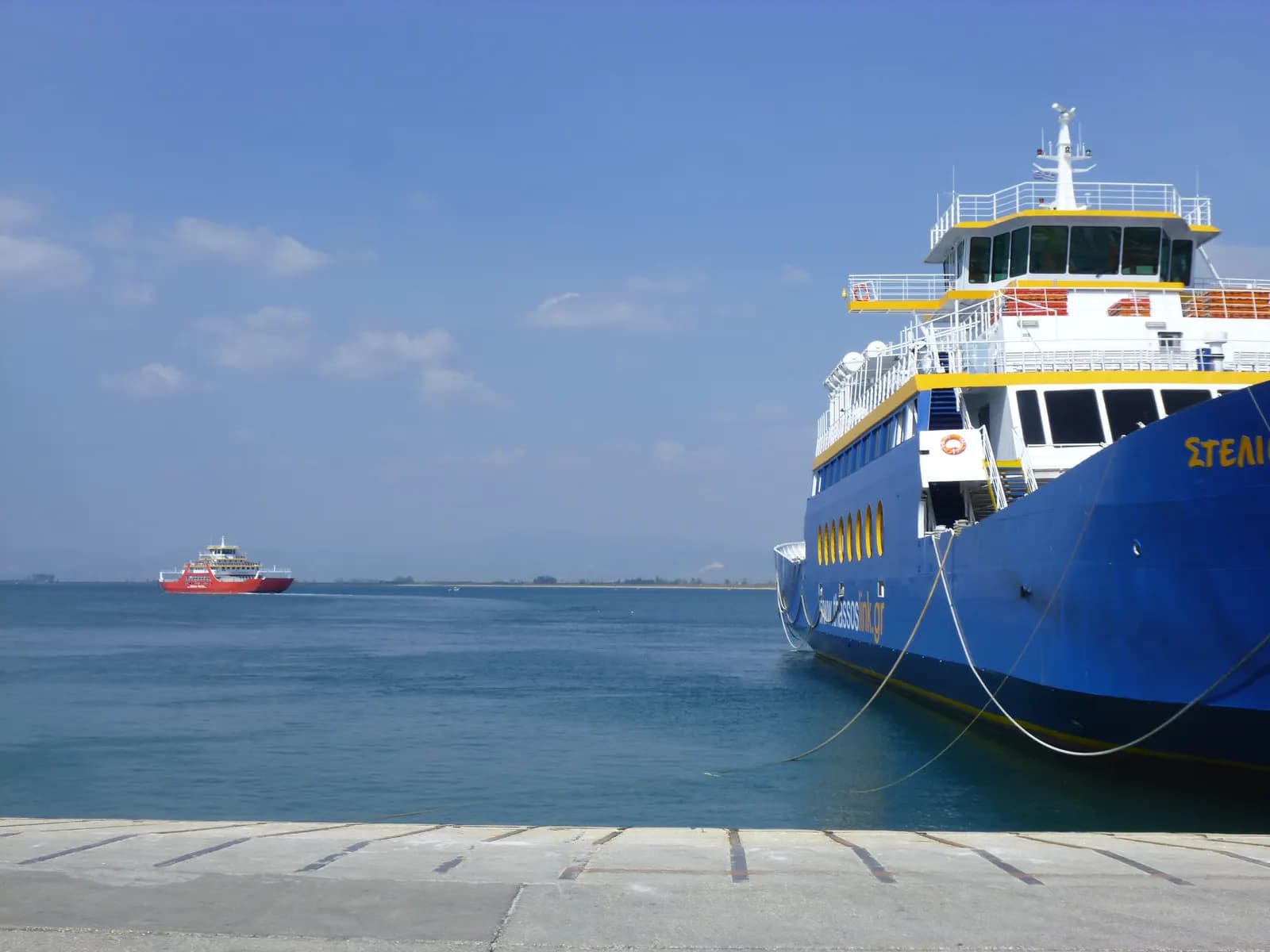 A blue and white car ferry moored at the quay in Keramoti, with a red ferry crossing the calm sea towards the port