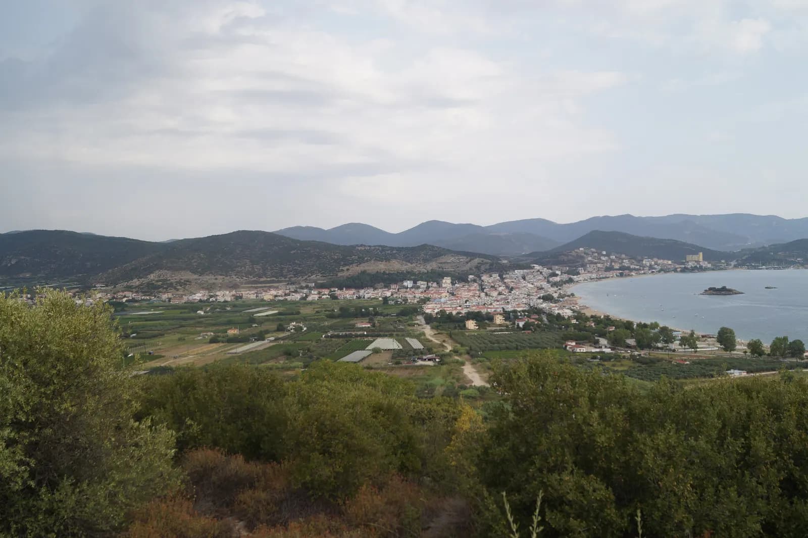 Nea Peramos seen from a hill, a coastal town on a curving bay with fields and greenhouses on the plain and wooded hills behind under a cloudy sky