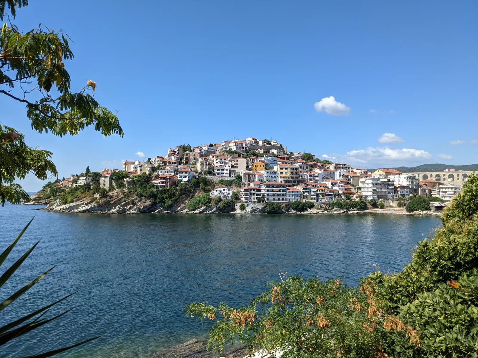 The Panagia peninsula of Kavala seen across the bay, its houses climbing a rocky headland above the water, with the arches of the aqueduct on the right