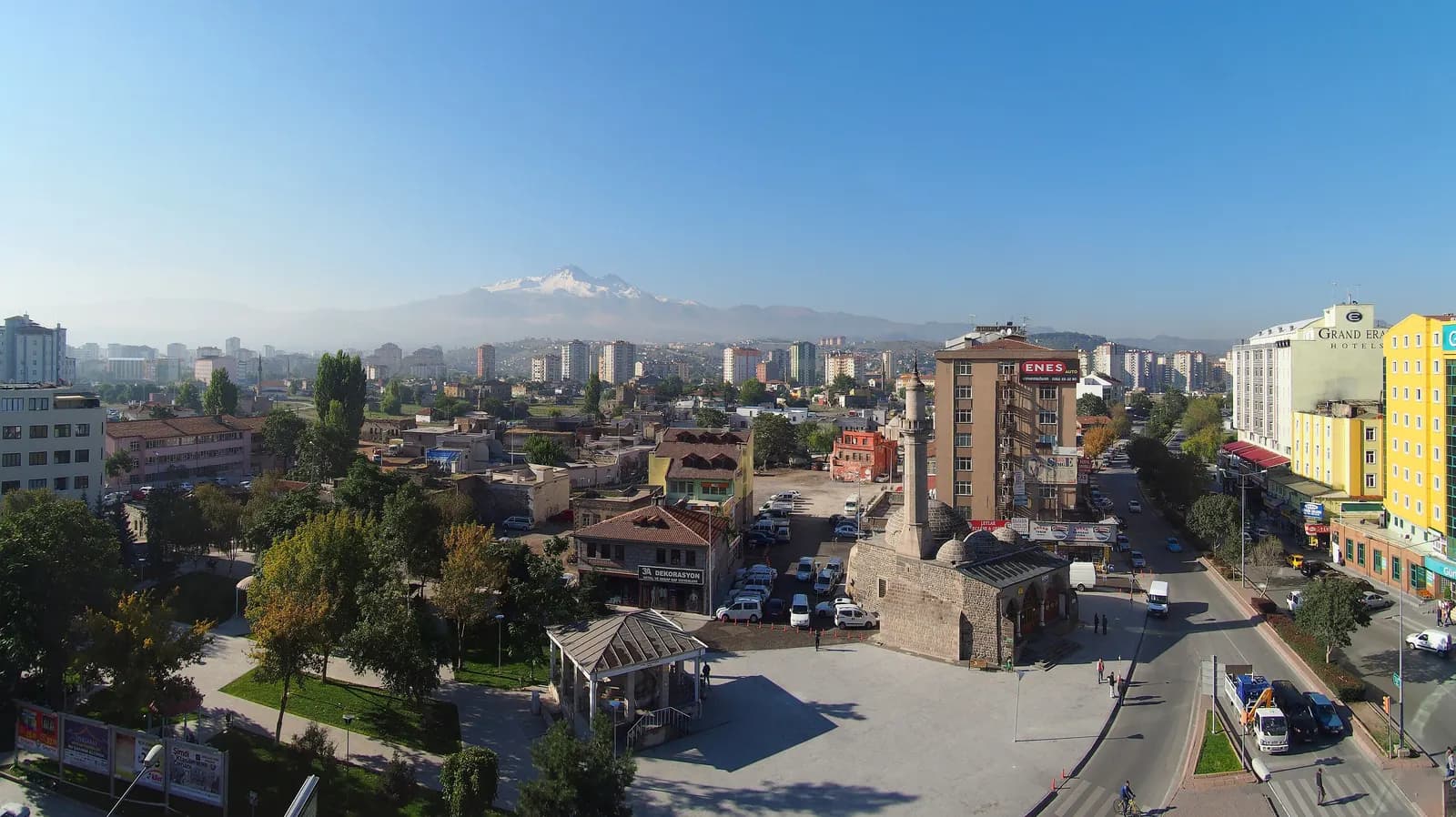 Central Kayseri from above, with a stone mosque and its minaret by a square and a car park, apartment blocks, and snow-capped Mount Erciyes on the horizon