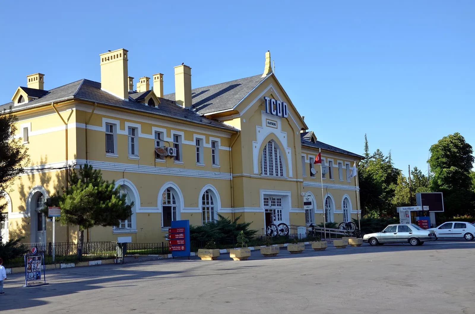 The yellow two-storey Kayseri railway station building with TCDD lettering on its gable, a forecourt and parked cars, under a clear sky