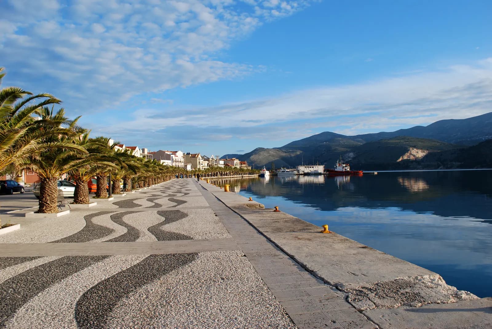 The waterfront promenade in Argostoli in early morning light, with a row of palm trees, patterned pebble paving, the harbour quay and mountains across the bay