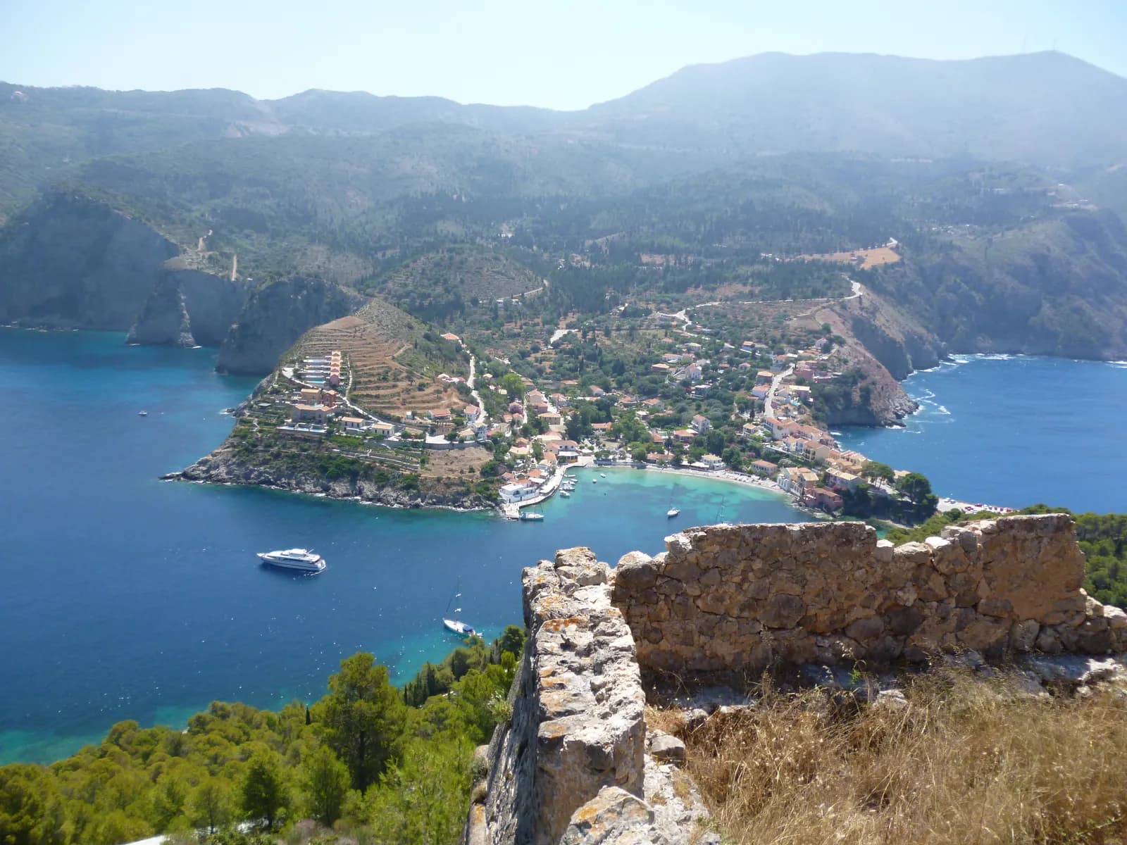 Assos seen from the walls of its Venetian castle, a village of pastel houses on a narrow neck of land between two bays, with boats at anchor and wooded mountains behind