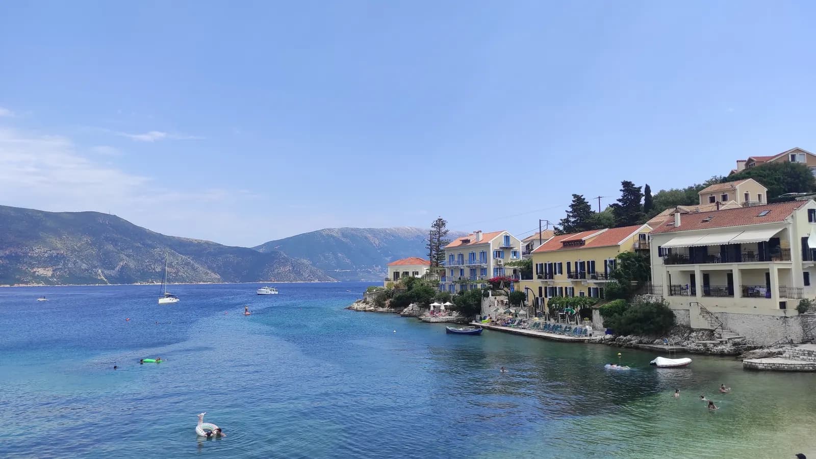 Pastel painted houses on the rocky shore at the entrance to Fiskardo harbour, with swimmers and moored boats in clear water and hills across the channel