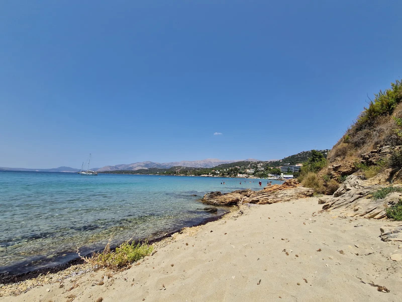 A sandy beach at Lassi with clear shallow water and flat rocks, swimmers further along the shore, hotels on the slope and mountains in the distance
