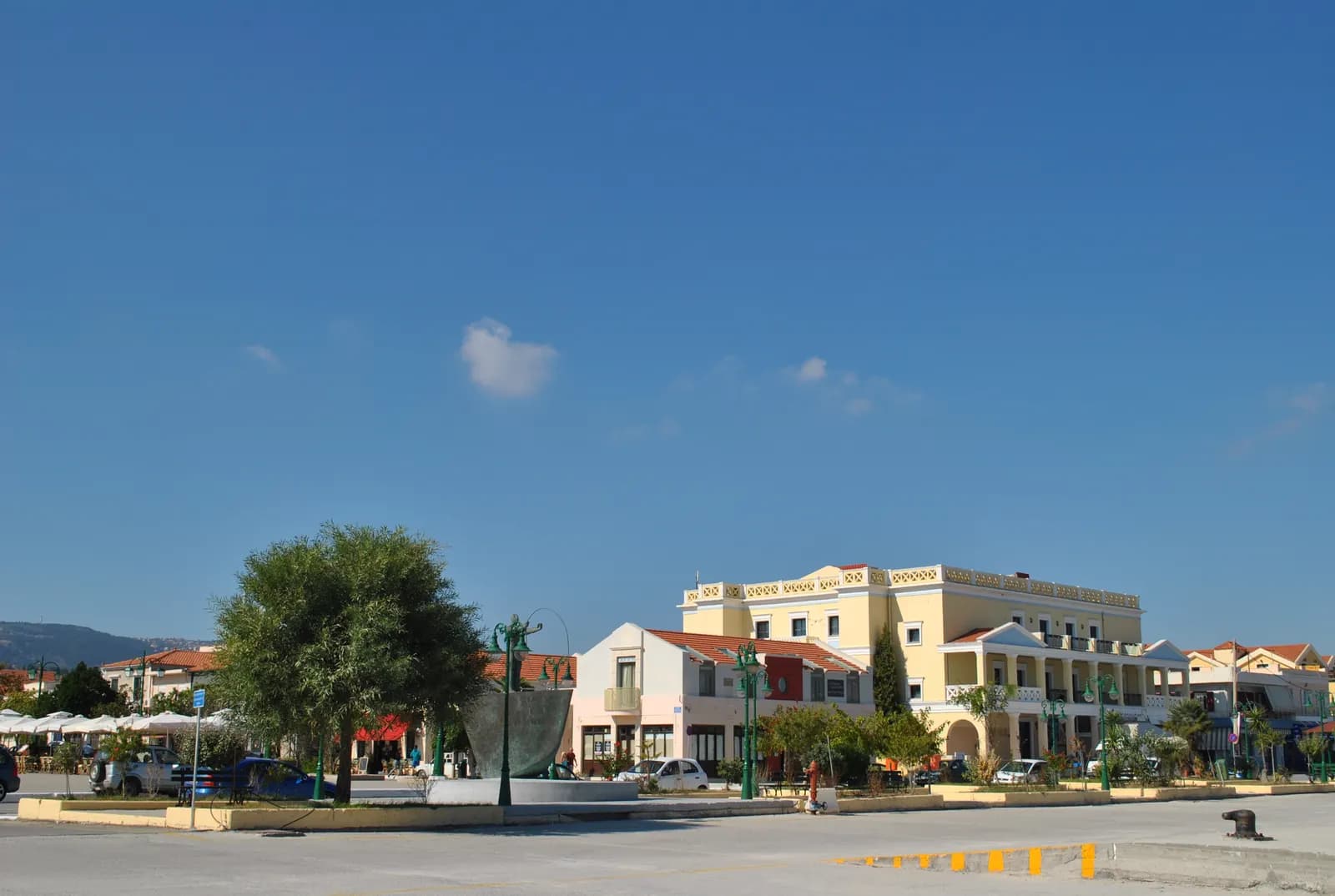 The main square of Lixouri seen from the quay, with cafe umbrellas, a bronze monument, a tree and a large pale yellow building with a columned porch