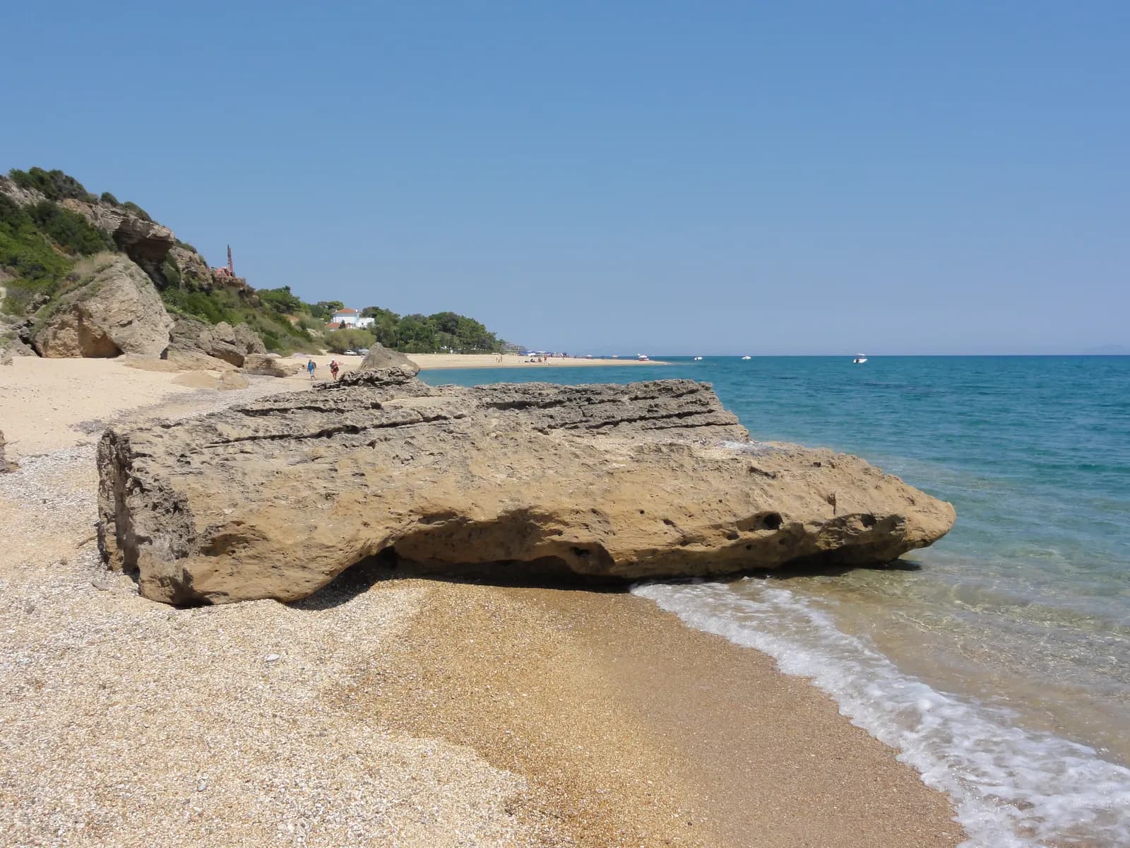 A large sandstone boulder at the water's edge on the beach at Skala, with clear turquoise water and the long sandy beach continuing below a wooded slope