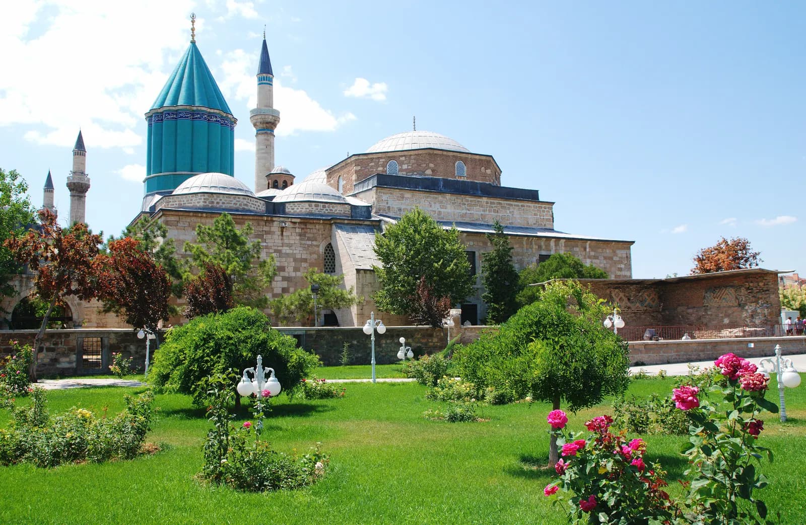 The Mevlana Museum in Konya with its turquoise conical dome and minarets, behind a garden of lawns, roses and trees