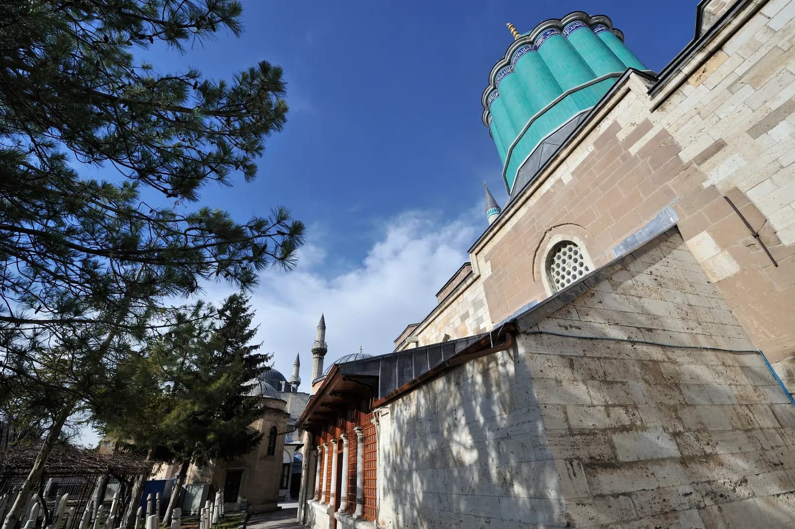 The fluted green dome of the Mevlana Museum above its stone walls, seen from the garden beside pine trees
