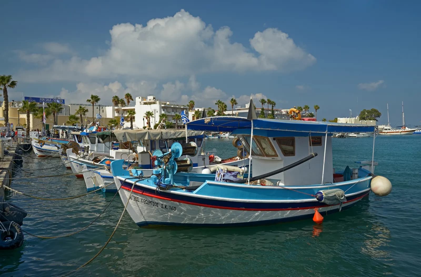 Blue and white fishing boats moored in Kos Town harbour, with palm trees and white buildings along the waterfront behind